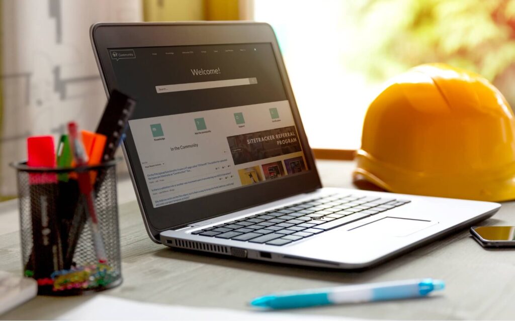 A laptop with Sitetracker Community on it, sitting on a desk with a yellow hard hat and pens