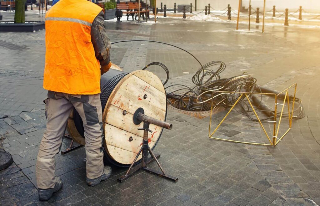 A fiber technician in a high-visibility jacket is handling a large spool of cables on a cobblestone street