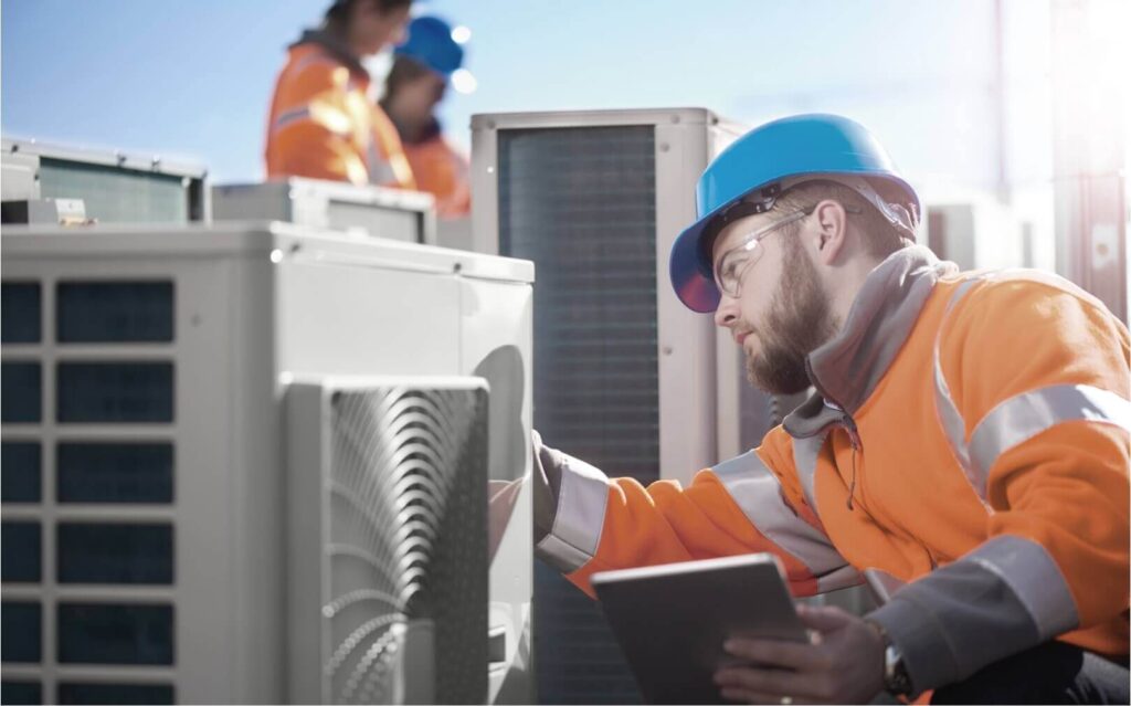 A worker in a blue hard hat and orange safety vest works on a HVAC unit while holding a tablet in one hand