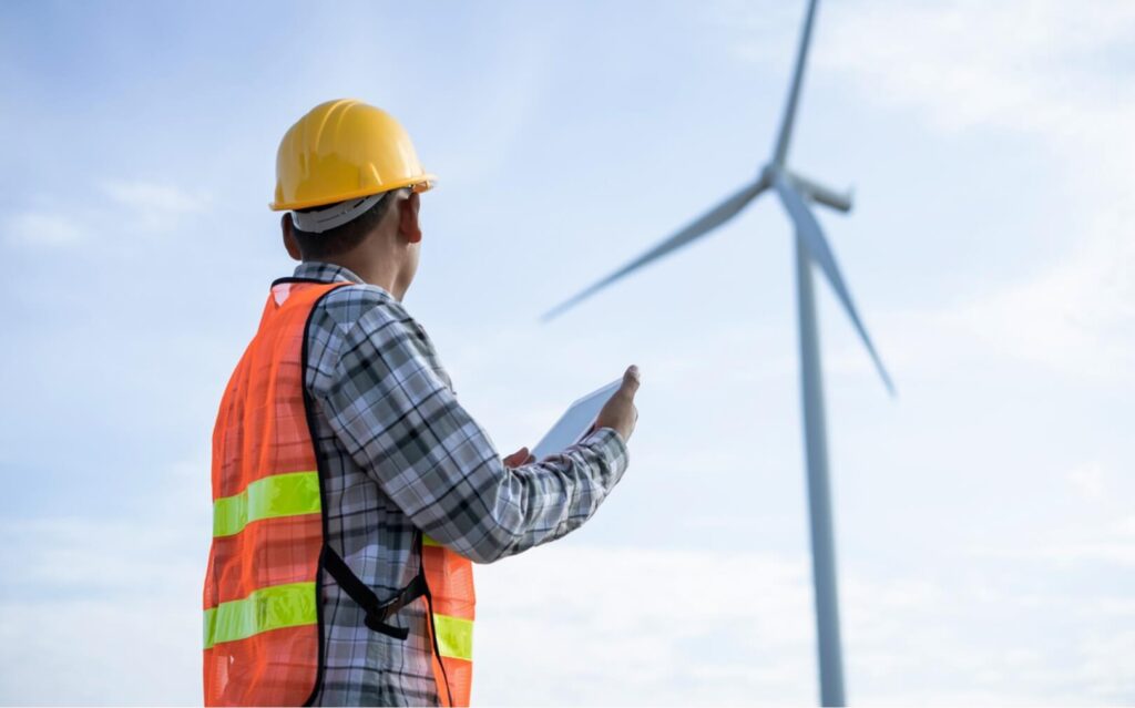 A field technician in a yellow hard hat and orange safety vest holds a tablet while looking up at a wind turbine in the distance 