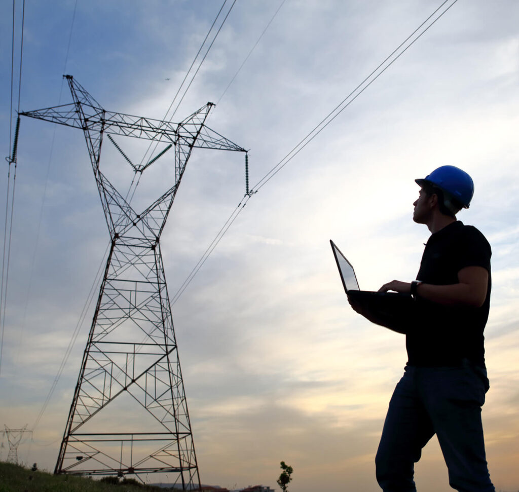 a field technician in a blue hard hat holding a laptop while looking up at an electrical tower in the background