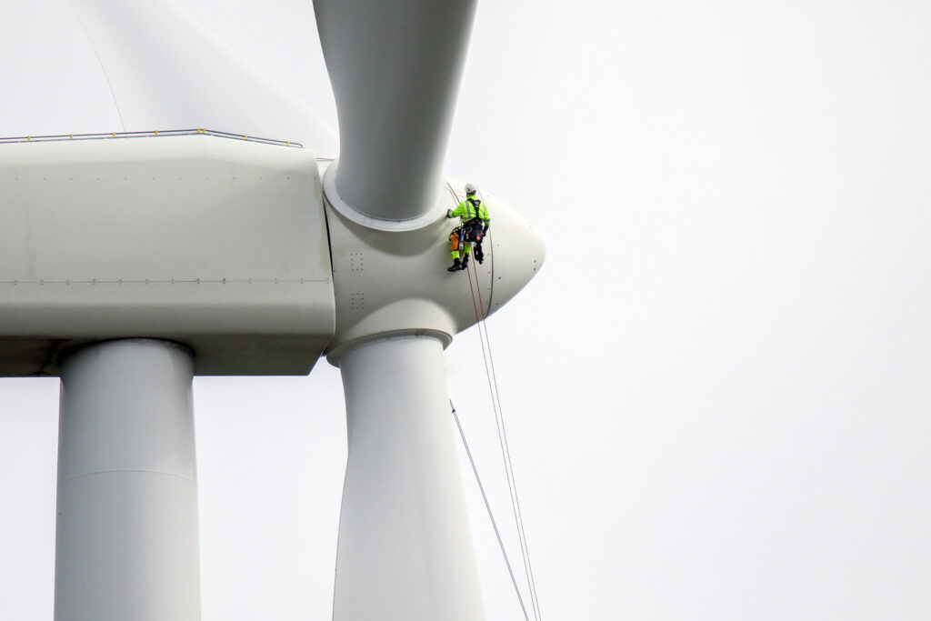 a technician in a white hardhat and yellow safety vest is suspended on the top of a wind turbine