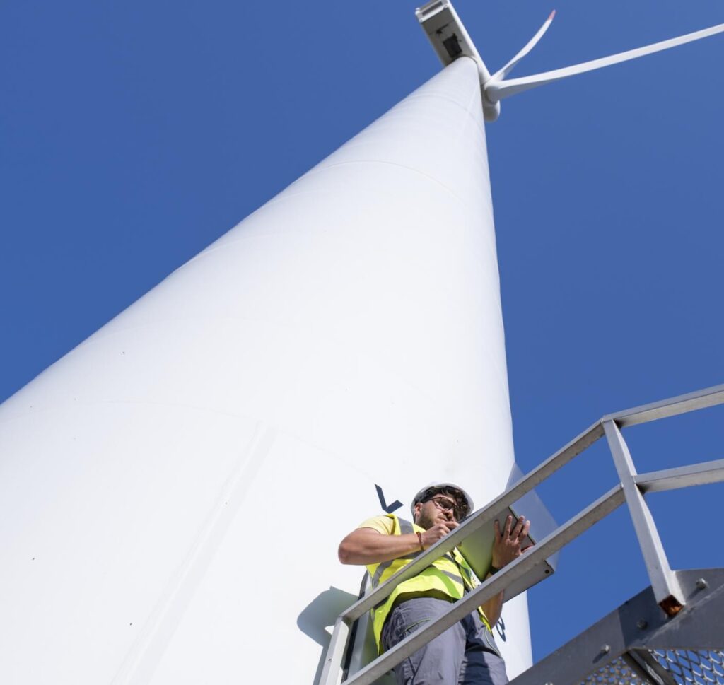A field technician in a white hard hat and yellow safety vest holding a laptop at the base of a wind turbine
