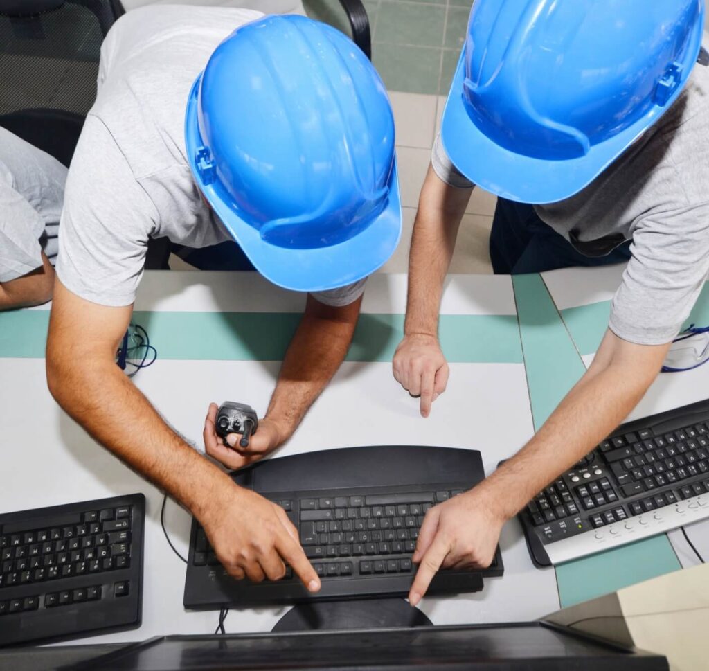 2 men in blue hardhats point to a computer monitor