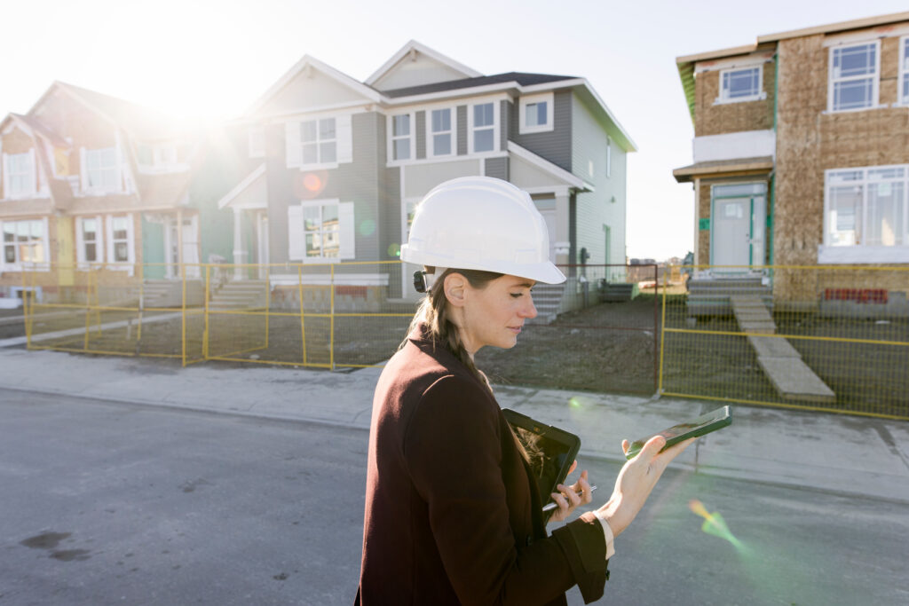 A woman in a hard hat holding a tablet and looking at a cellphone walks past a row of houses under construction