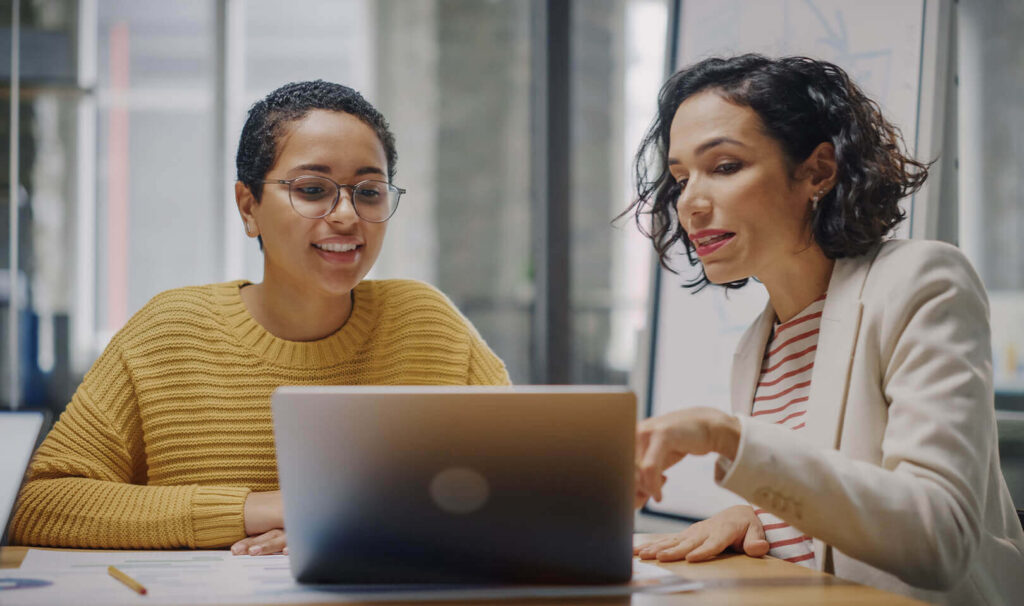 2 female professionals collaborate on a laptop in an office setting