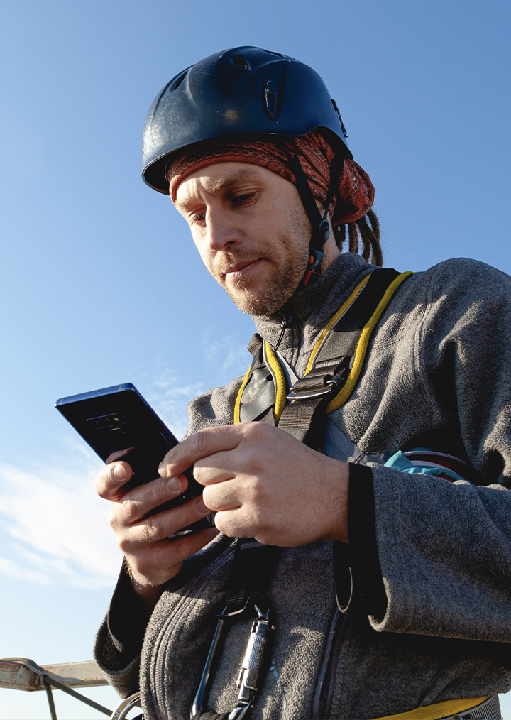 A field worker in a black hard hat and safety gear looks at his cell phone