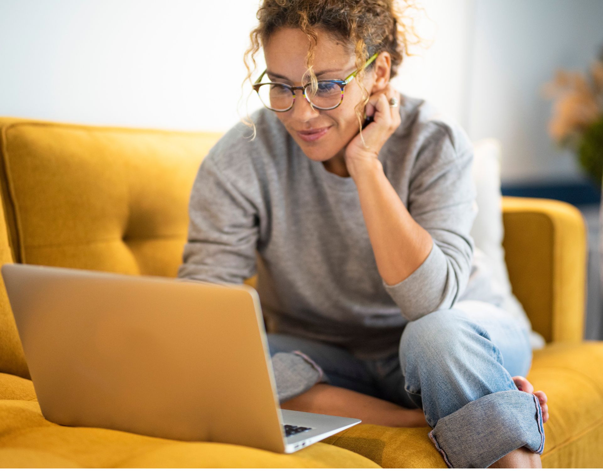 A woman with glasses casually sitting on a mustard yellow couch working on her laptop and smiling