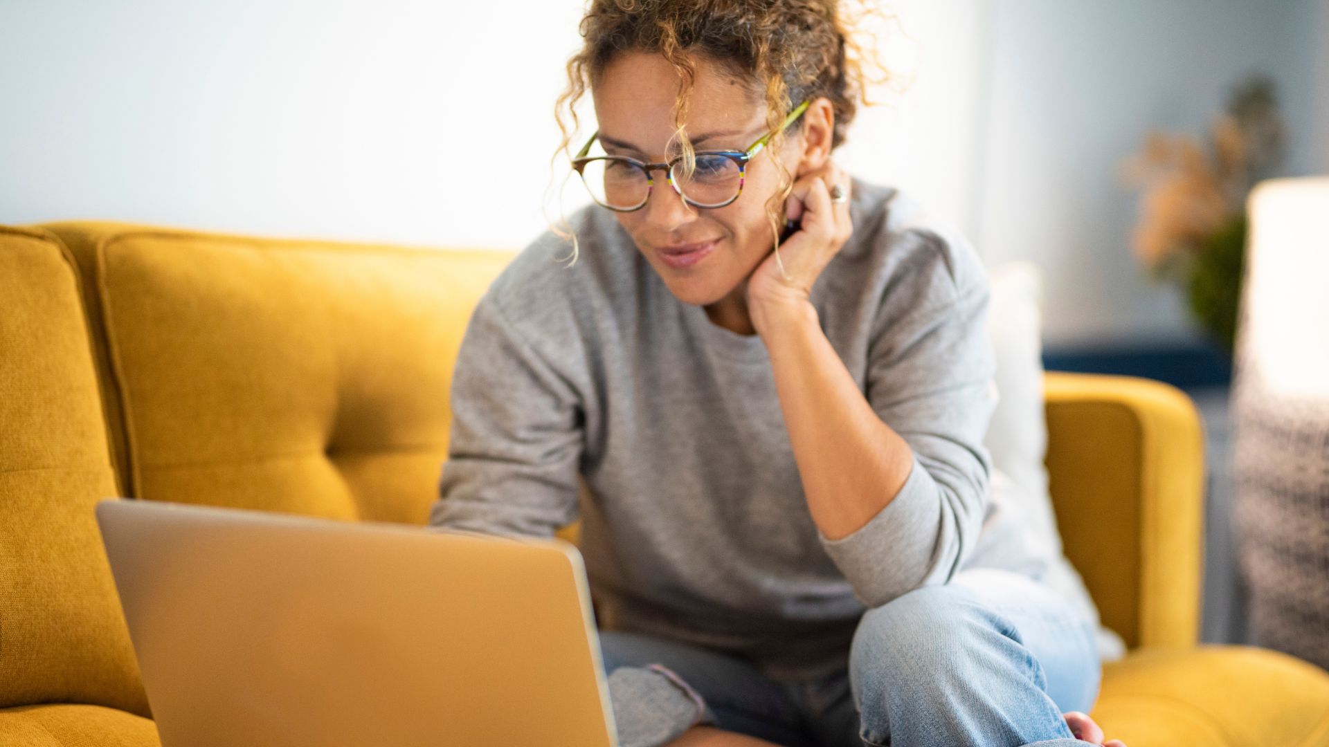 A woman with glasses casually sitting on a mustard yellow couch working on her laptop and smiling