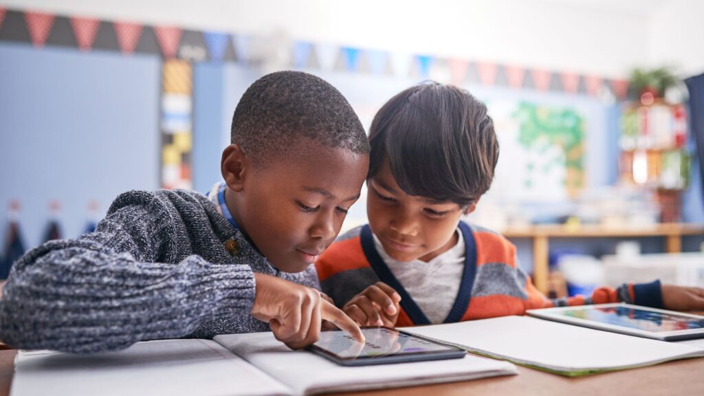 Two young boys in a classroom interacting with a tablet