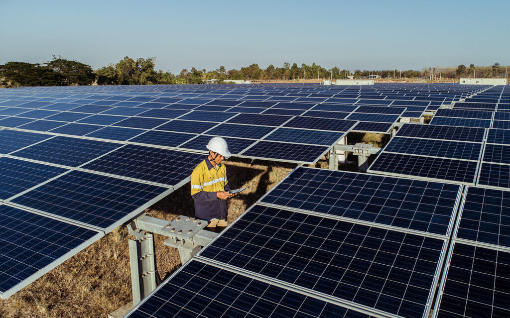 A field technician in a white helmet looks at a tablet while standing in a field of solar panels