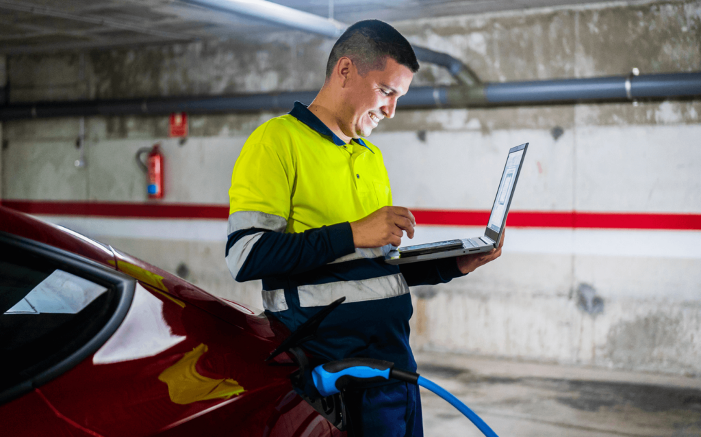 An EV contractor leaning on a charging electric vehicle while smiling and working on a laptop