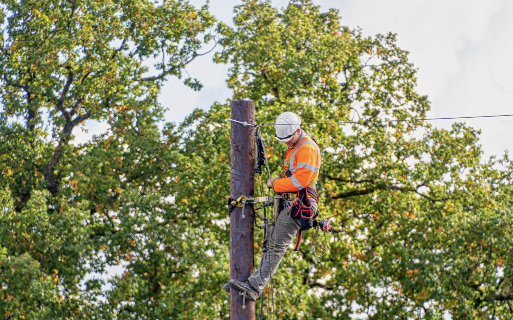 A fiber contractor in an orange safety vest and white helmet works atop a utility pole