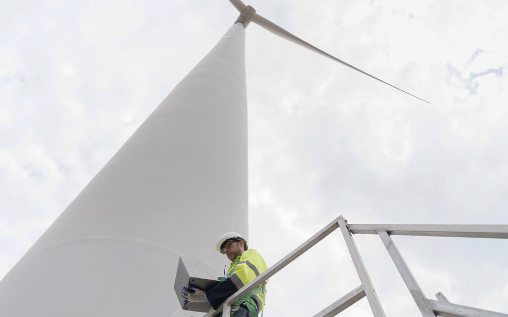 A wind turbine technician wearing a yellow safety vest and white helmet works on his laptop while standing at the base of a wind turbine