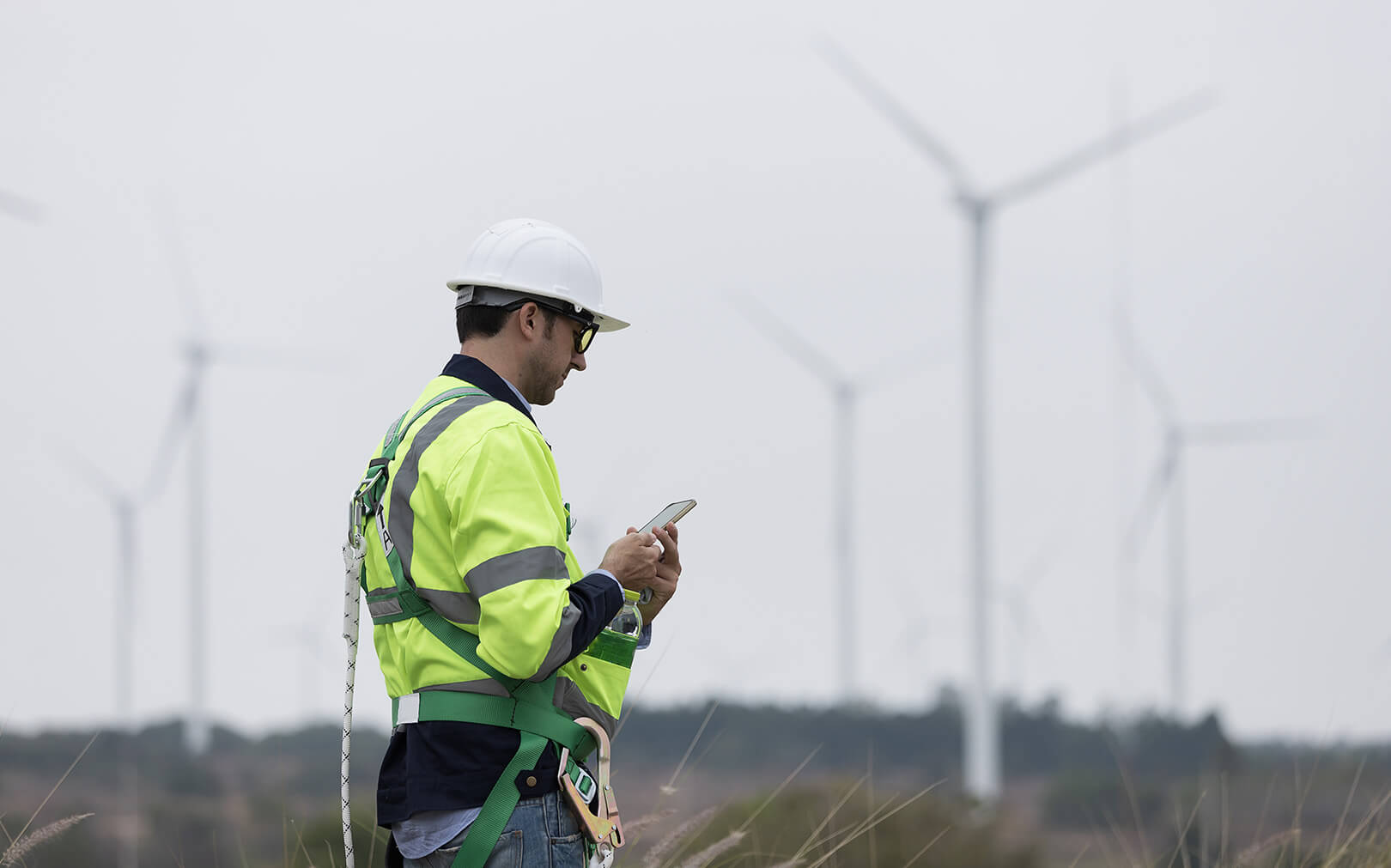 A field technician in a white helmet and safety gear looks at his cellphone with wind turbines in the distance