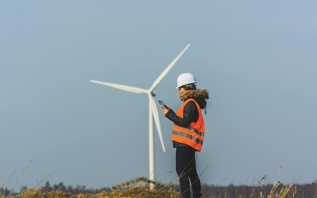 A female field technician in an orange safety vest and white helmet looks at a tablet in her hands with a wind turbine in the background