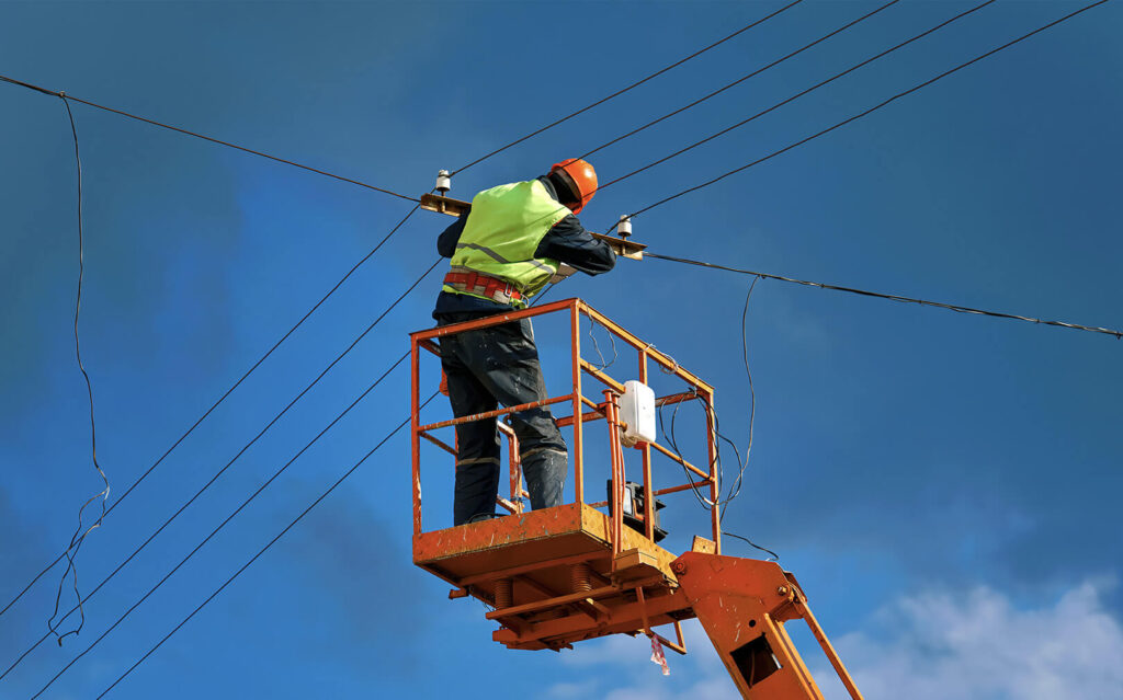 A worker in a high-visibility vest and safety helmet is standing on an elevated platform repairing electrical lines against a clear blue sky