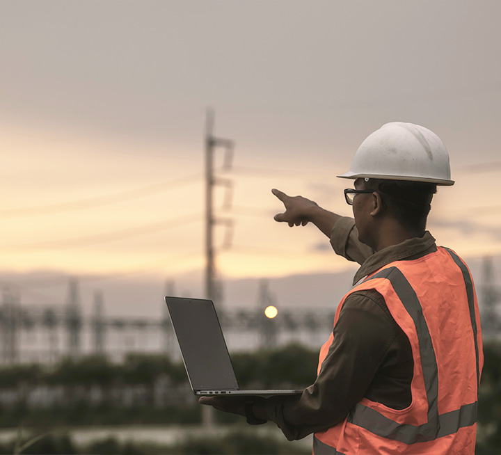 A worker in safety gear holding a laptop points his finger to something in the distance