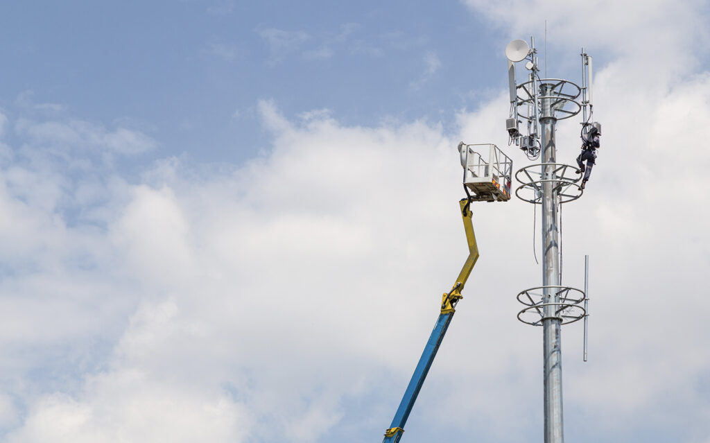 Technician in a harness working on a cell tower, assisted by a crane with a blue arm against a cloudy sky