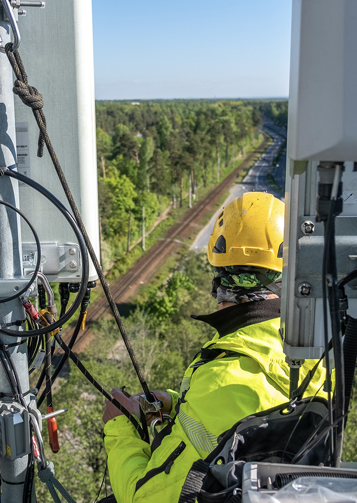 A technician in a yellow helmet and high-visibility jacket is working on telecommunications equipment high above the ground with a forest and railway track in the background