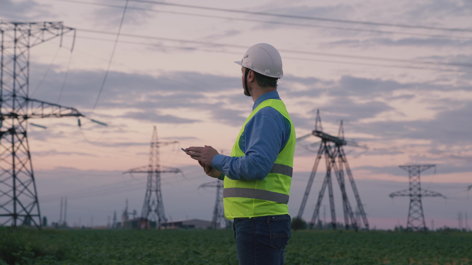An engineer in a reflective vest and helmet stands in a field at dusk, holding a tablet with high-voltage power lines and pylons in the background.