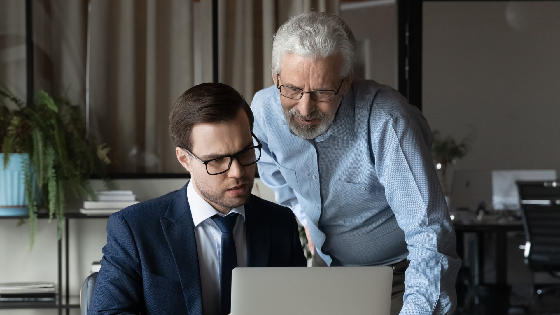 An older man mentoring a younger colleague while working together on a laptop in an office setting.