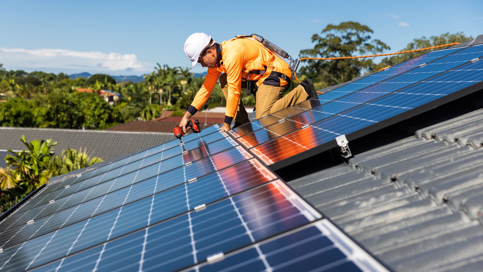A construction worker in a bright orange safety jacket and white hard hat is installing solar panels on a residential rooftop