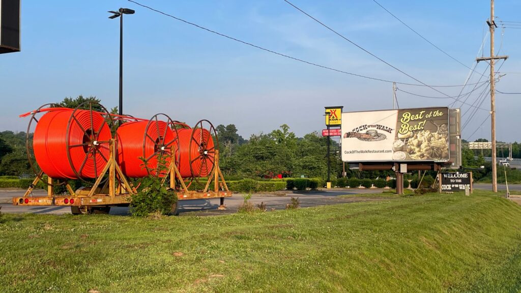 Three large orange fiber optic cable reels are mounted on a trailer parked beside a grassy roadside, with a "Cock of the Walk" restaurant billboard and KOA campground sign visible in the background.