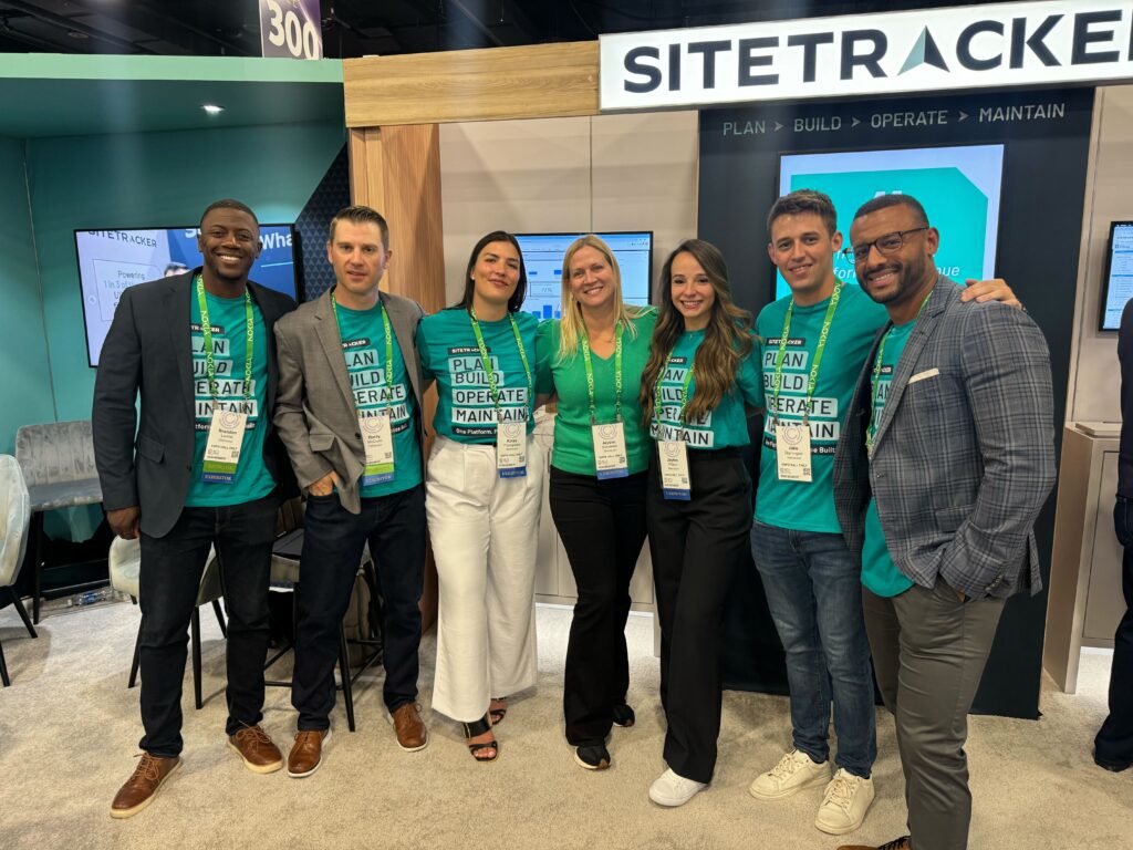 A group of seven Sitetracker employees stands together smiling in front of a conference booth, wearing matching teal t-shirts with the slogan "Plan Build Operate Maintain" and name badges.