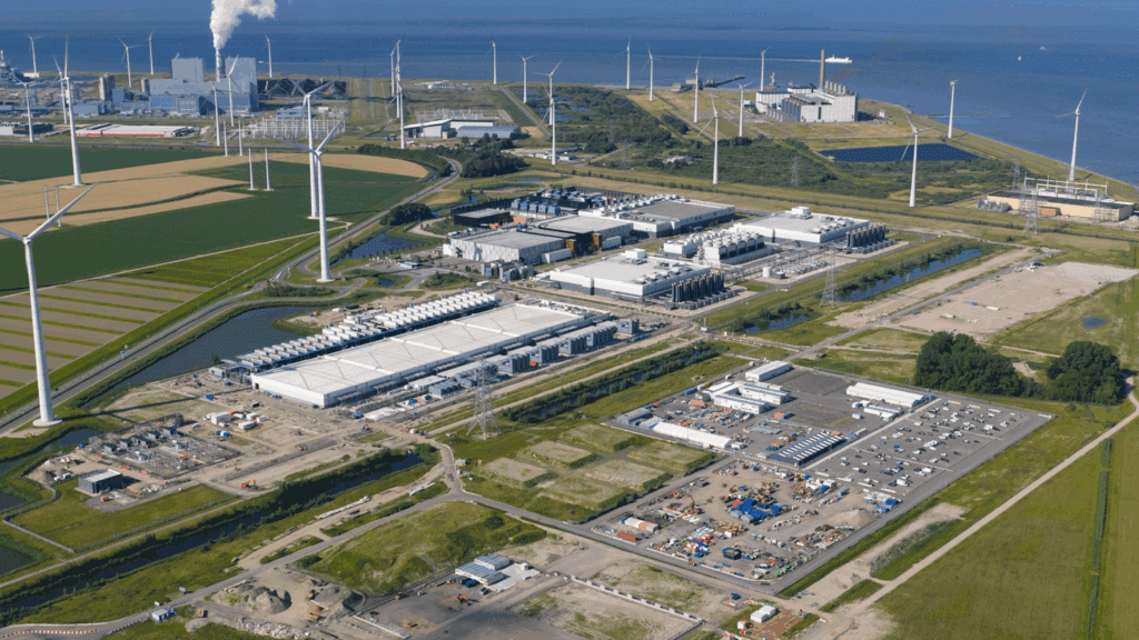 Aerial view of a large data center campus near the coast, surrounded by farmland and wind turbines. Multiple large white-roofed buildings, construction areas, parking lots, and utility infrastructure are visible, with a power plant and smokestack in the background.