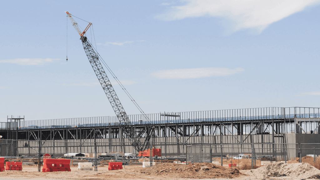 Construction site of a data center with a large crane lifting materials. The steel framework of the building is visible, with workers and vehicles operating in the background.
