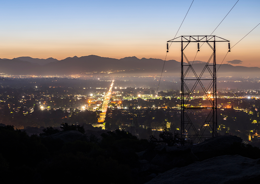 A twilight view of a city stretching into the distance, illuminated by streetlights and building lights. In the foreground, a tall electrical transmission tower stands against the backdrop of mountains and a fading orange-blue sky