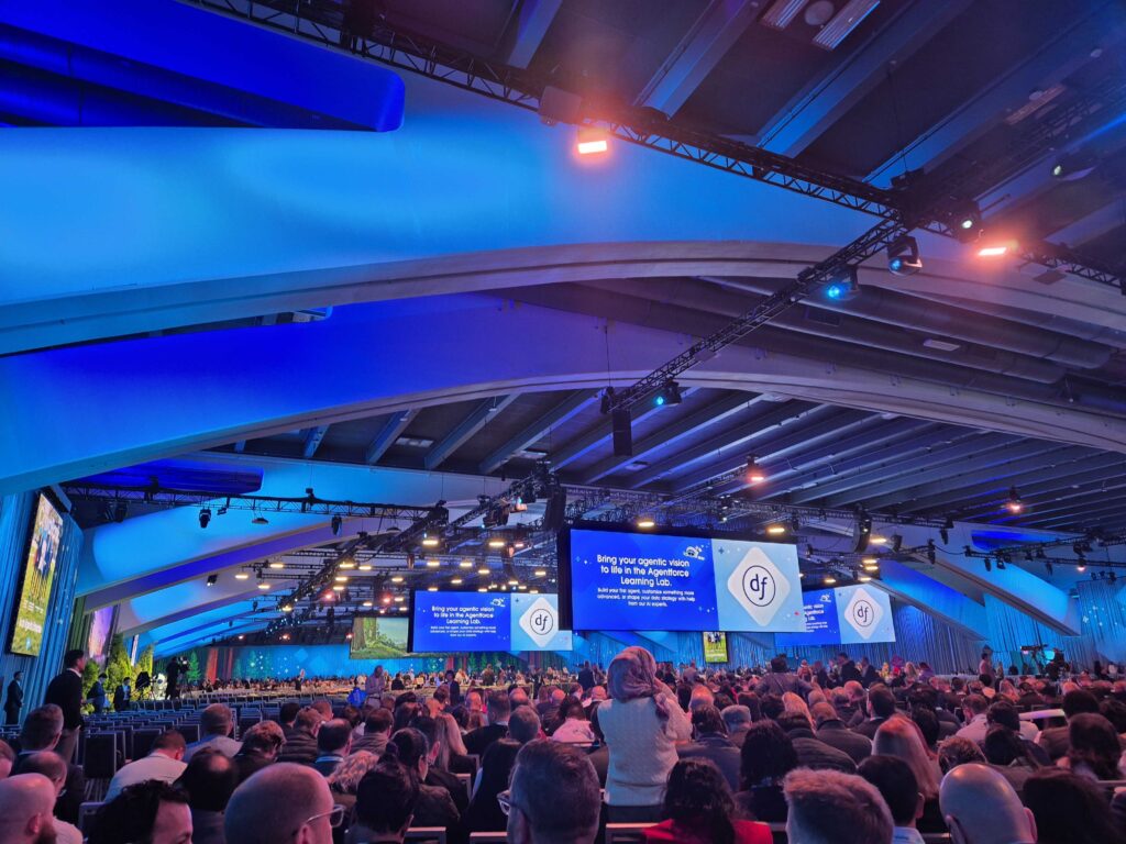 Large crowd seated inside a blue-lit convention hall with curved ceiling beams, multiple large display screens showing the Salesforce Dreamforce logo, and a presentation in progress.