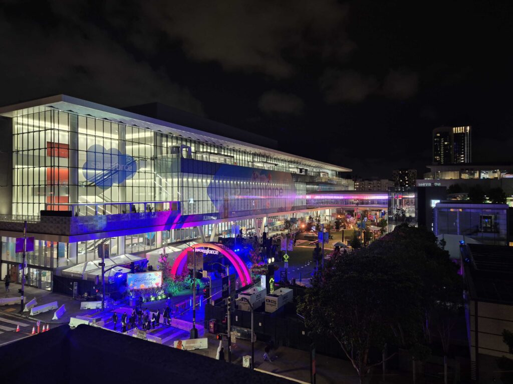Nighttime view of the Moscone Center in San Francisco illuminated with colorful pink, purple, and blue lights and large Salesforce logos during Dreamforce, with people walking outside below.