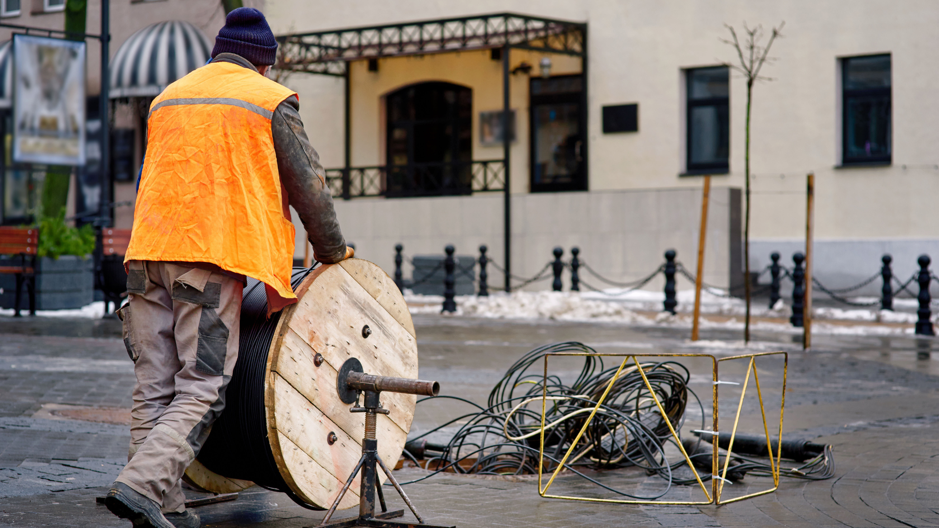 A construction worker in an orange safety vest unrolling a large wooden spool of fiber-optic cable on a city street, with tools and cable scattered nearby