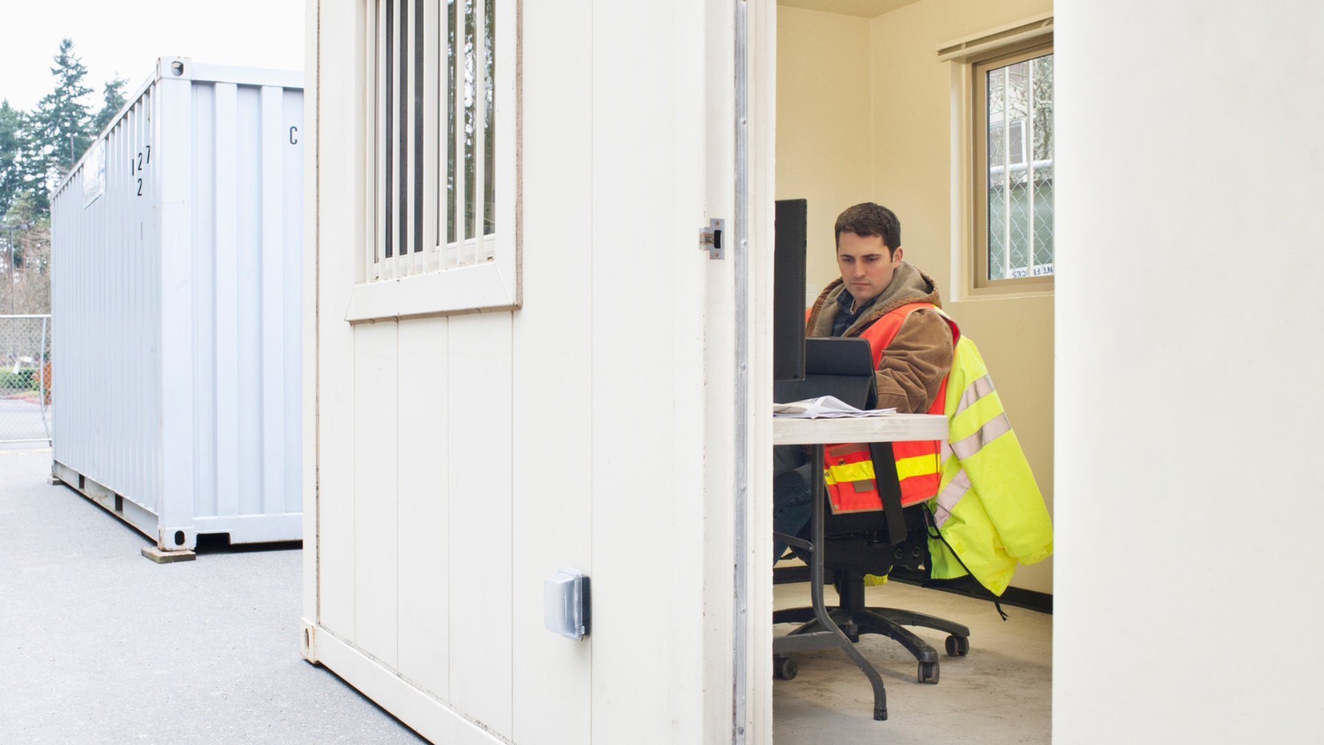 A worker wearing a reflective safety vest sitting at a desk inside a portable job-site office, reviewing documents and working on a computer