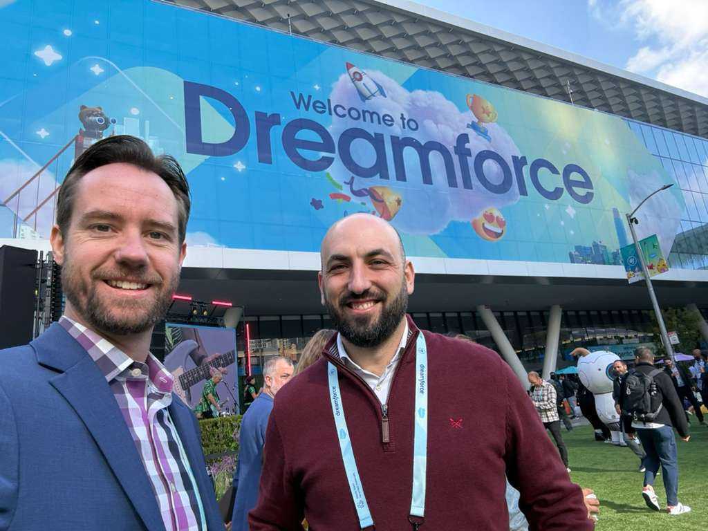 Two smiling men posing in front of a large ‘Welcome to Dreamforce’ banner on the Moscone Center exterior, surrounded by attendees and outdoor exhibits under daylight