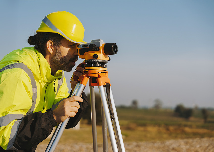 Field engineer in a hard hat using a surveying level on a tripod at an outdoor job site