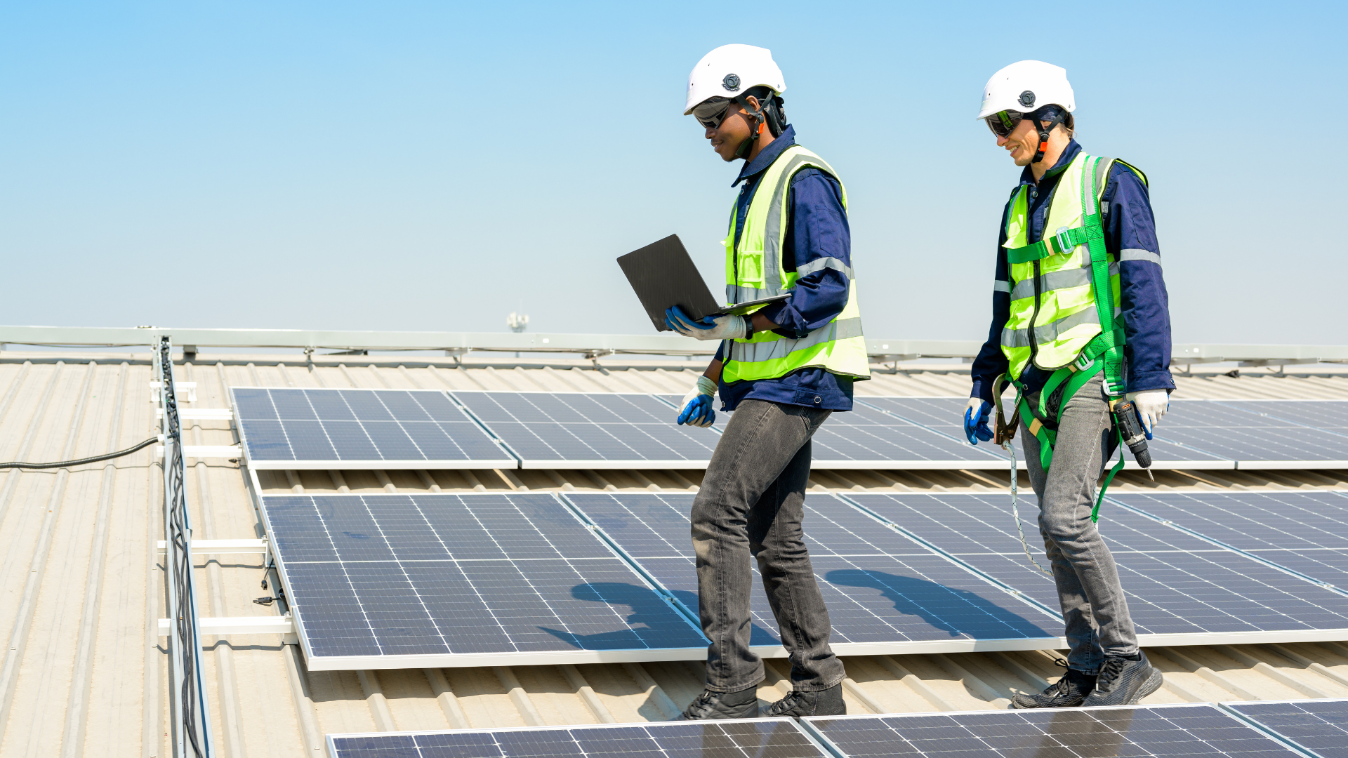 Two solar technicians wearing safety helmets and high-visibility vests walk on a rooftop covered with solar panels. One technician holds a laptop while the other carries tools, both inspecting the solar installation under a clear blue sky