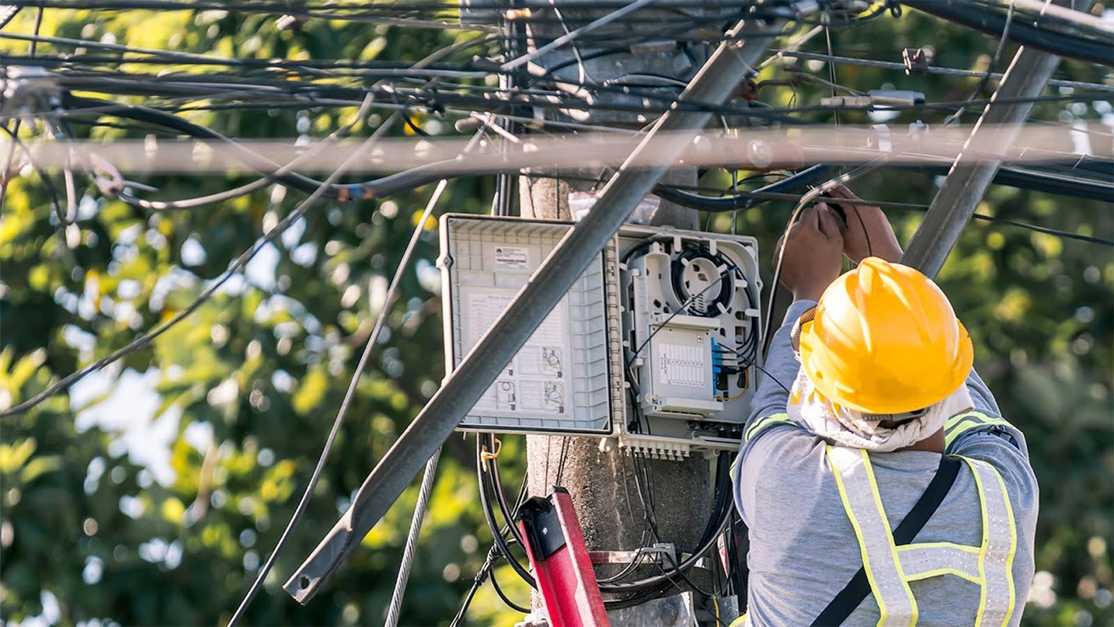 Field technician wearing a safety helmet and high-visibility vest performing maintenance on a fiber optic network box mounted on a utility pole
