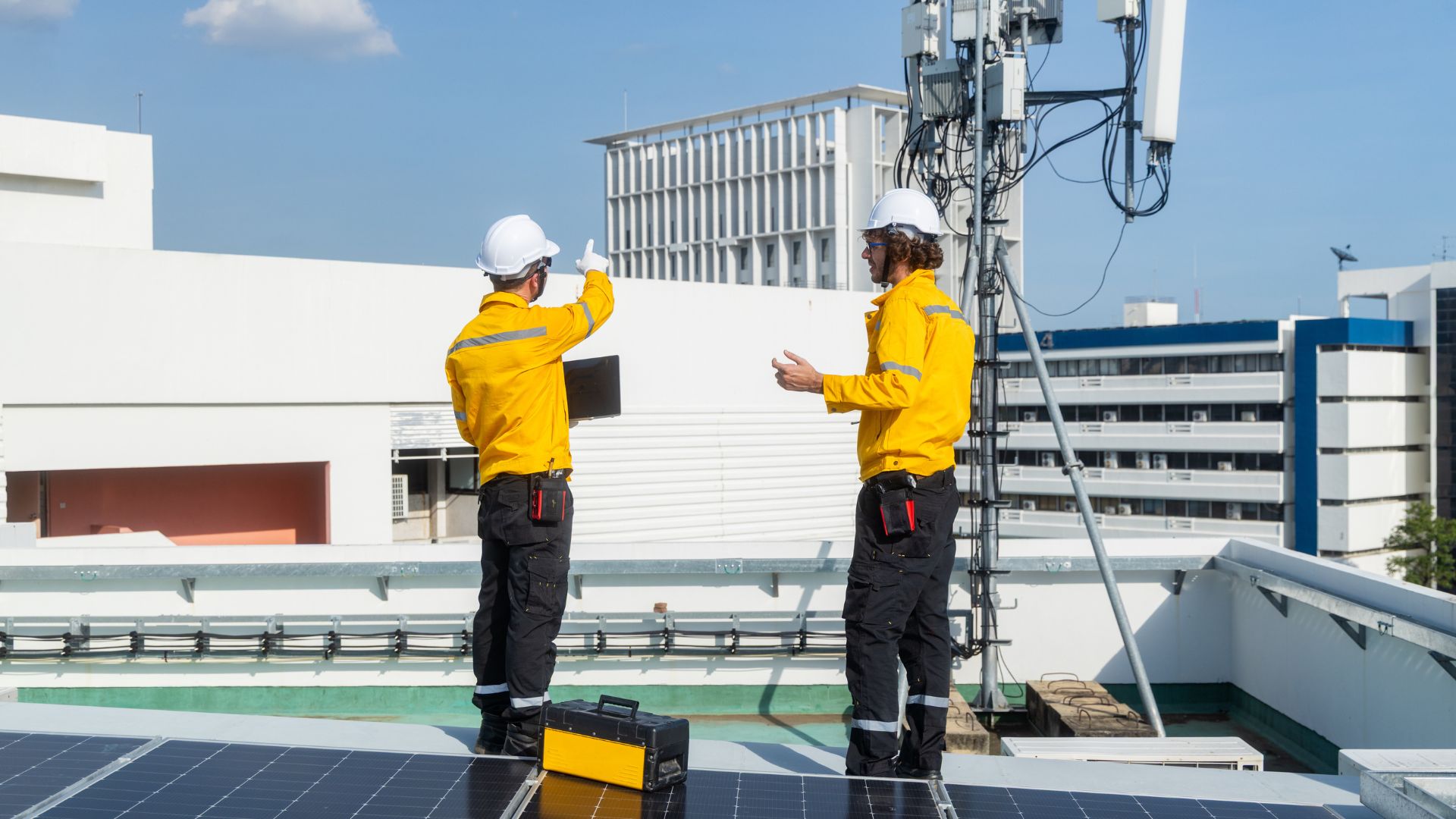 Two technicians in yellow safety jackets and white hard hats stand on a rooftop near telecom antenna equipment and solar panels, with one pointing toward nearby buildings in the background