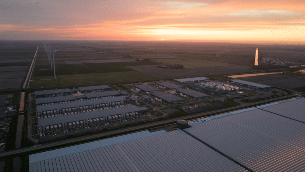 Aerial view of a large data center campus at sunset, with rows of data centers and cooling units in the foreground, expansive agricultural fields surrounding the site, and wind turbines and canals stretching into the distance under a colorful orange and pink sky.
