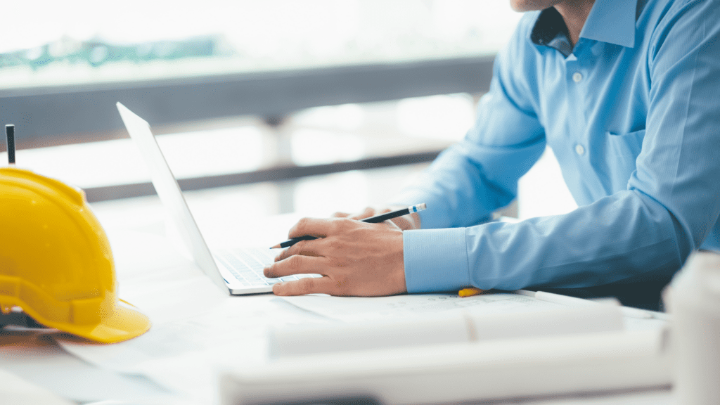 Person in a blue shirt working on a laptop while holding a pencil at a desk with construction plans and a yellow hard hat nearby, suggesting project management