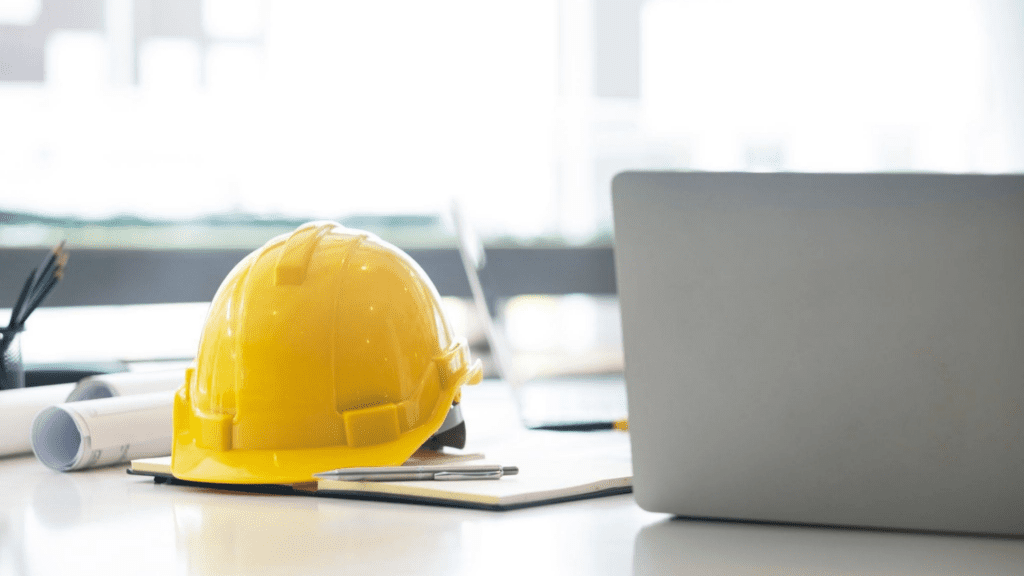 A yellow construction hard hat resting on a desk beside a notebook and pen, with a laptop and rolled-up plans in the background, suggesting an engineering or construction workspace