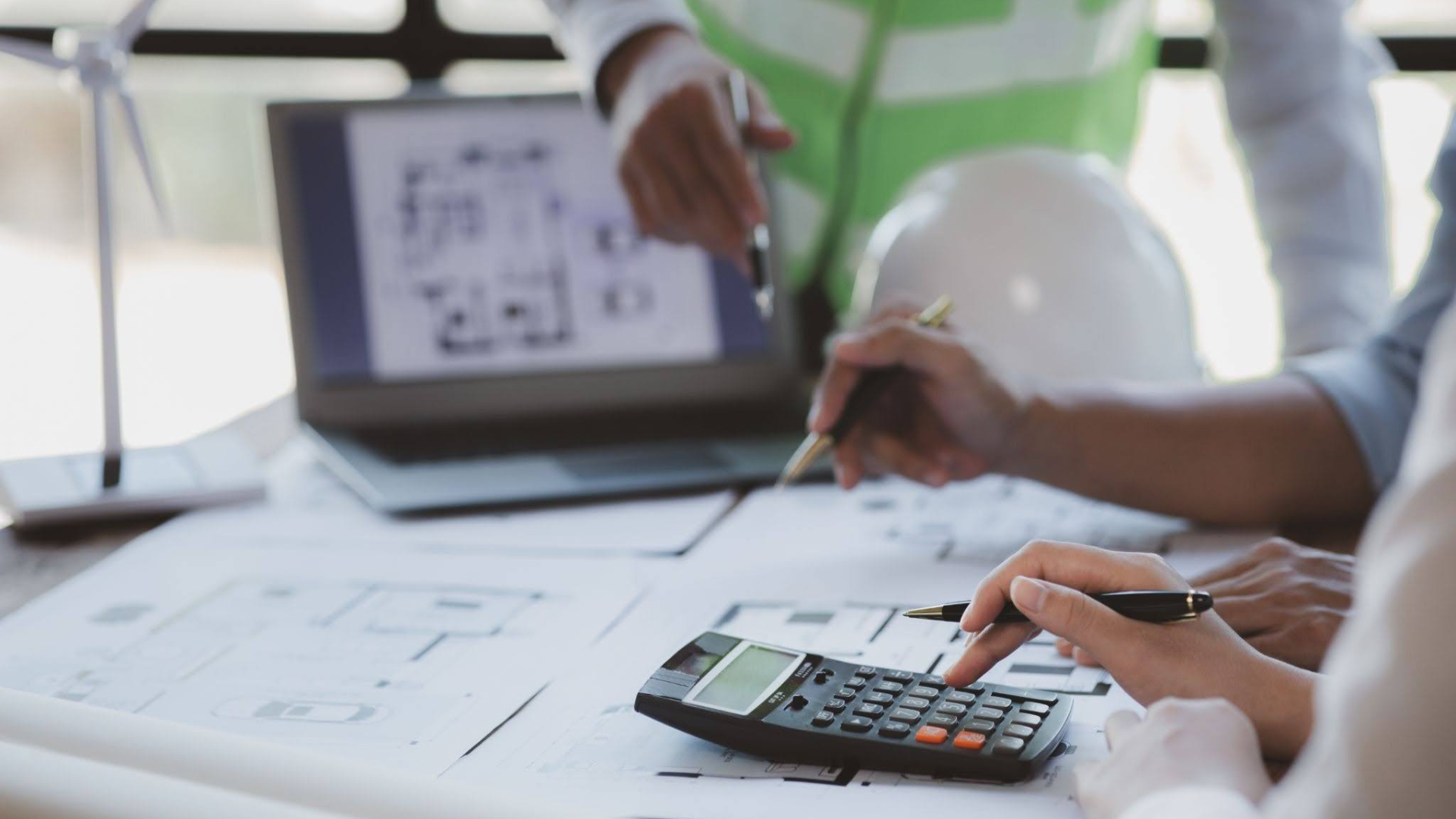 Two professionals reviewing construction plans at a desk, using a calculator and laptop, with a hard hat and wind turbine model visible, suggesting construction or engineering project planning and budgeting