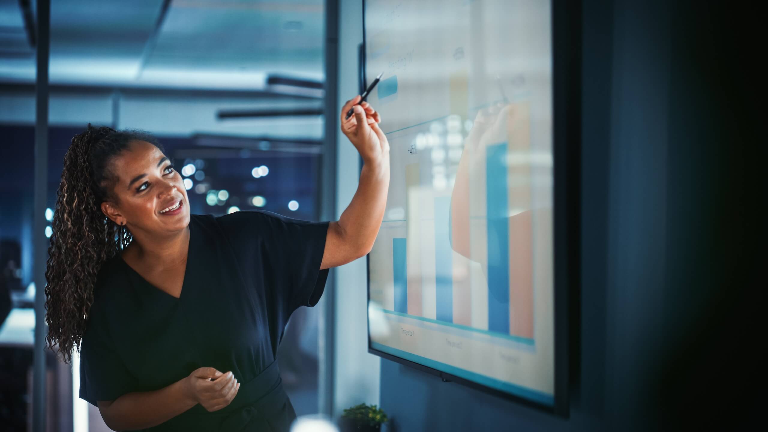 Woman presenting data on a large screen, pointing at a bar chart in a modern office setting