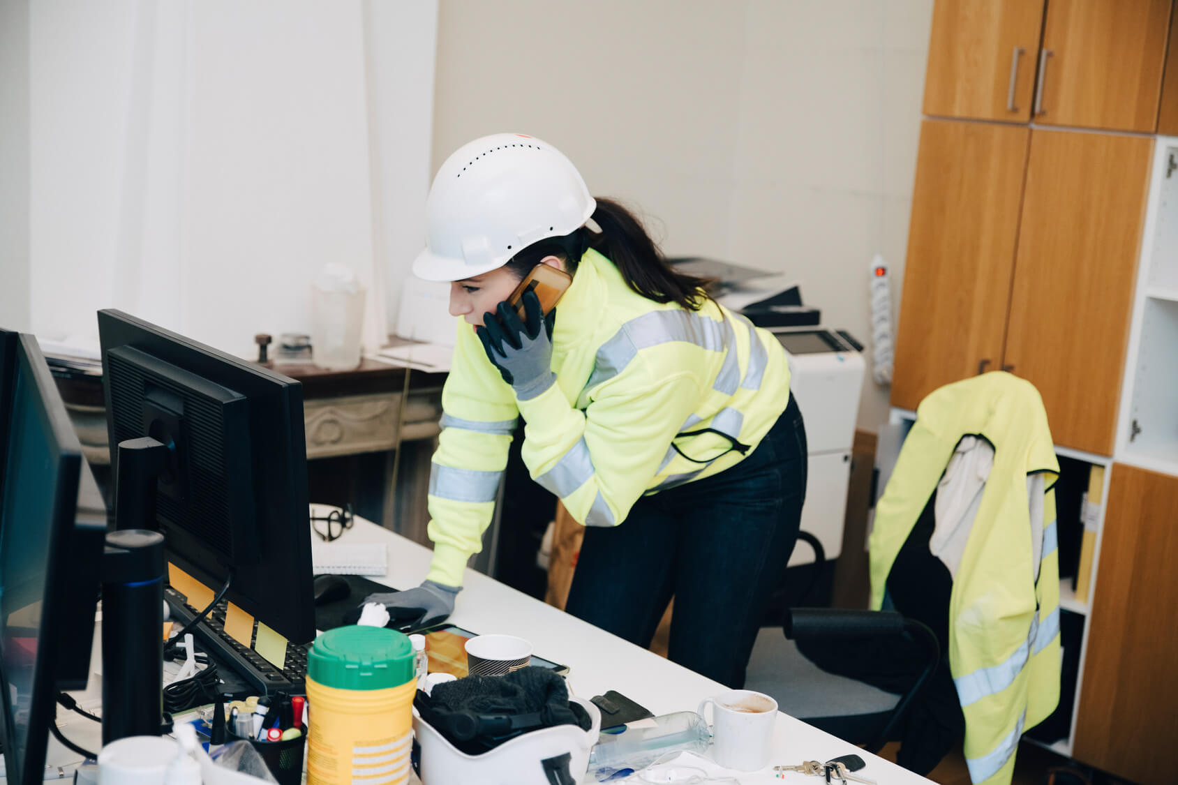 Construction worker wearing a white hard hat and high-visibility jacket talking on a phone while leaning over a desk with computer monitors