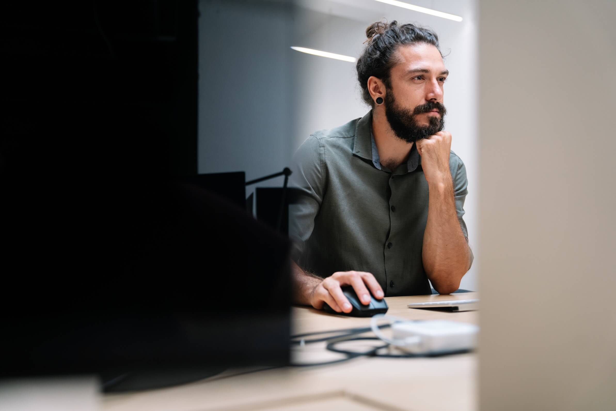 Man with long hair and beard working at a computer in a modern office, resting his chin on his hand while using a mouse