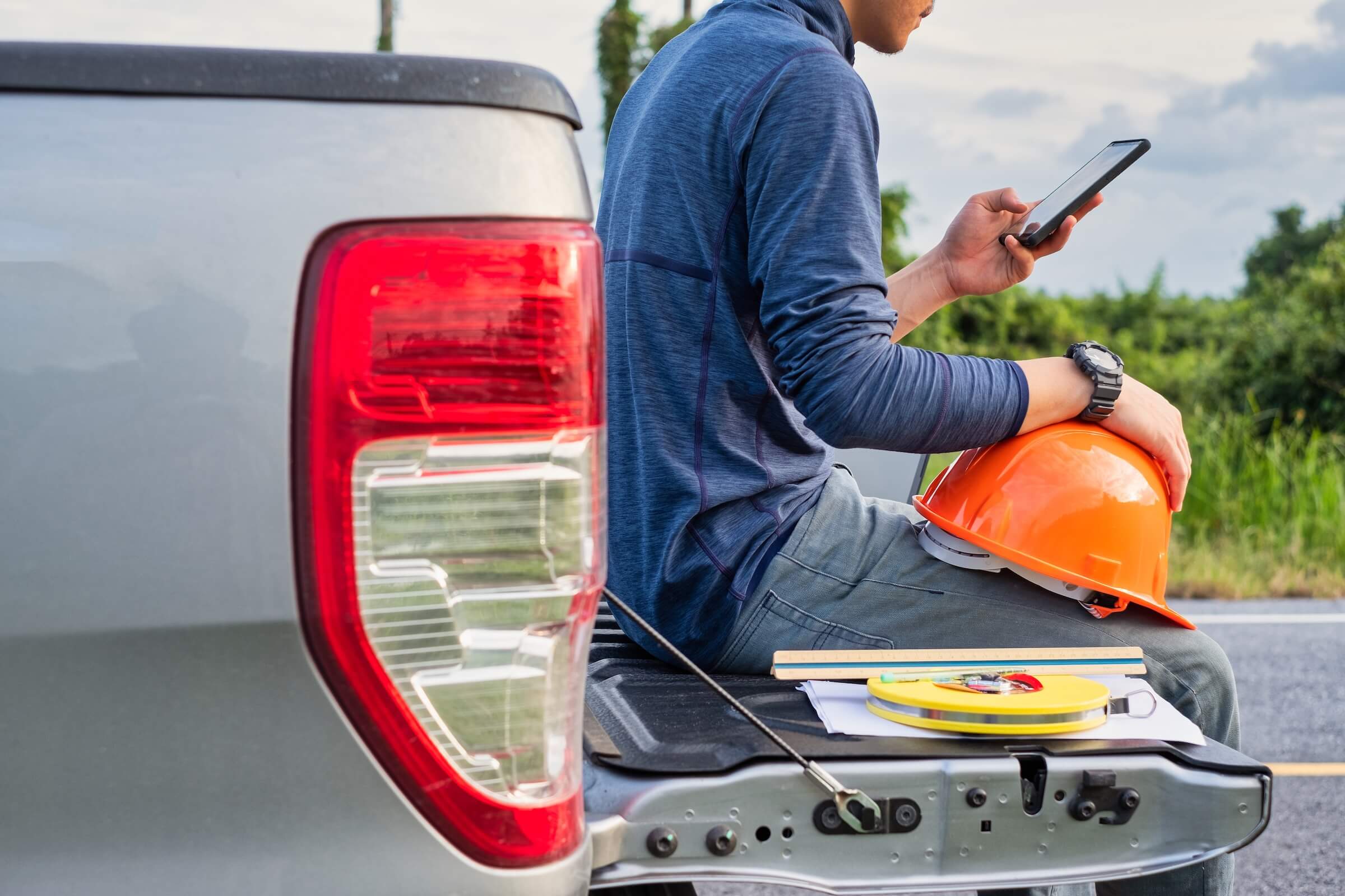 Technician sitting on the back of a pickup truck using a smartphone, with a hard hat and tools beside him