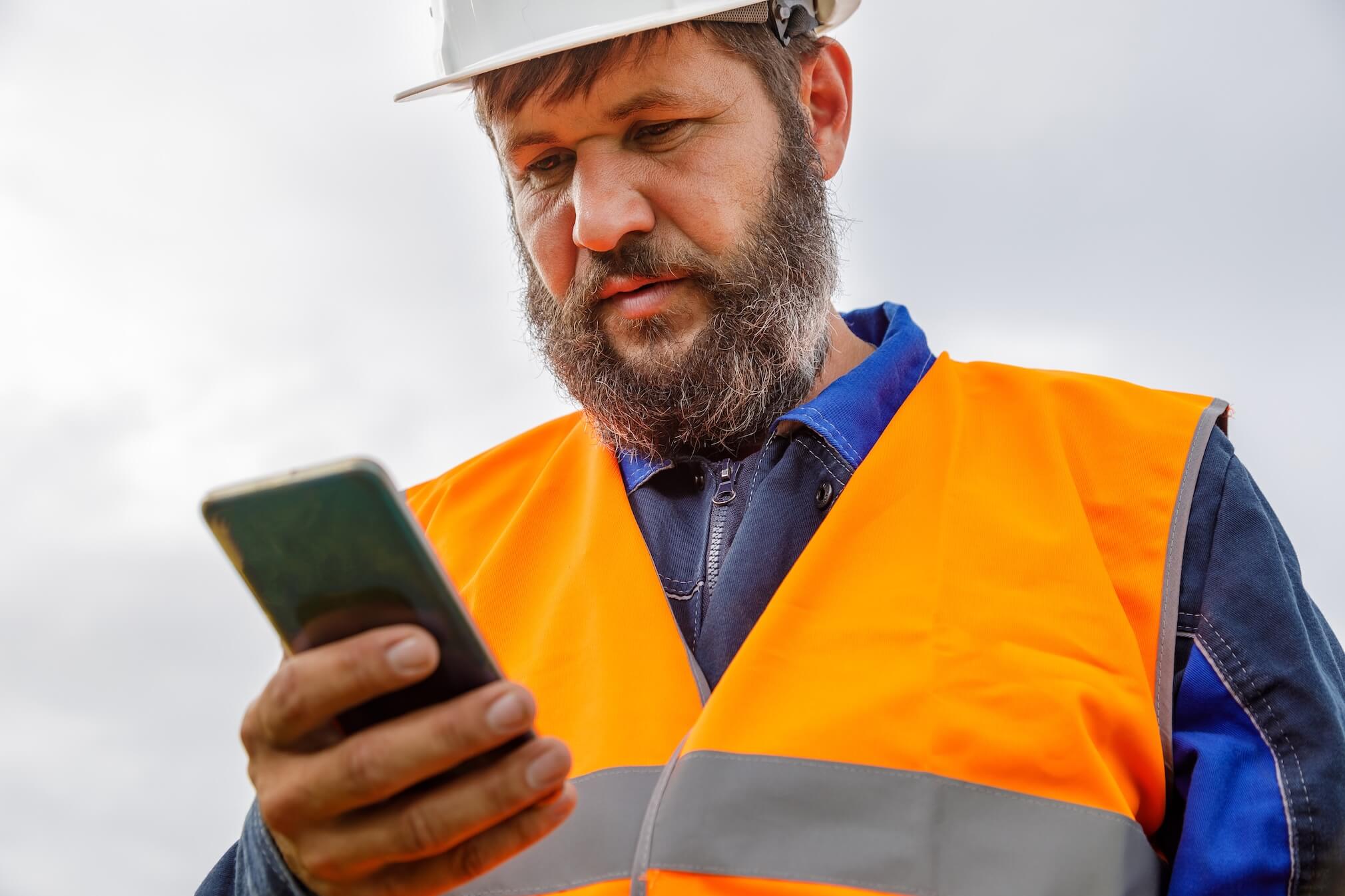 Bearded worker in an orange safety vest and hard hat looking at a smartphone